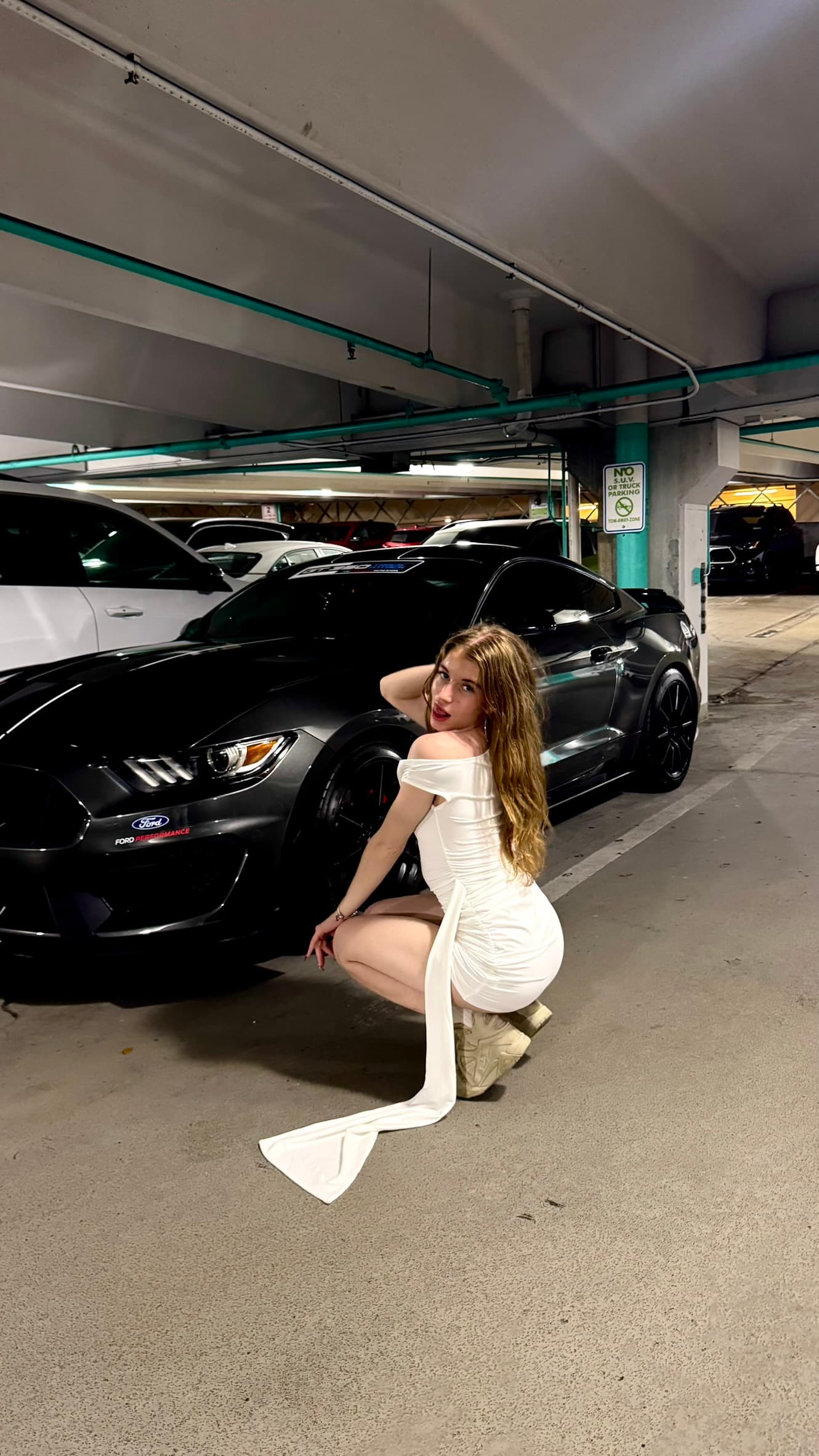 Mia Doll in a white gown beside a black sports car